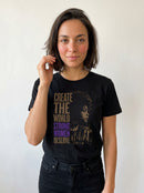 gravisk Create The World Strong Women Deserve organic graphic tee. A woman's profile beside a city skyline, gold type with STRONG WOMEN in purple.
